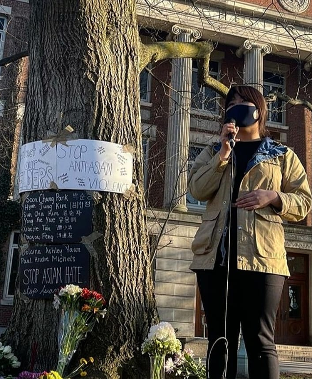 A person wearing a face mask holding a microphone in front of a tree

Description automatically generated